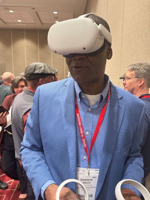 Mr. Alford is looking up in a suit wearing a VR headset and using the handheld controllers.